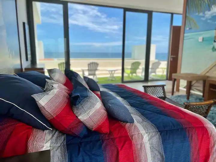 Cozy bed with red, blue, and white bedding by a large window overlooking the beach.