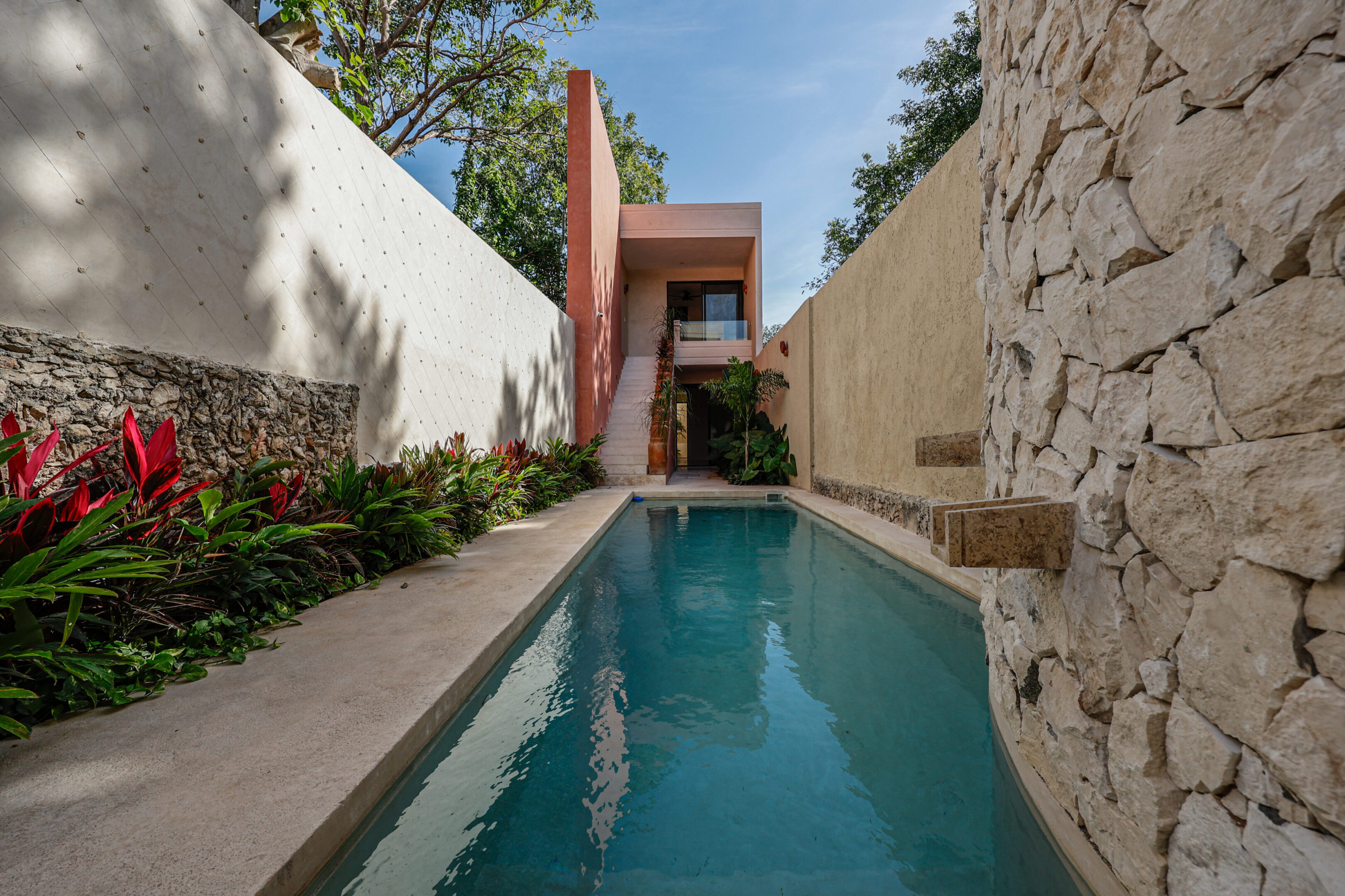 A narrow pool between stone and white walls with plants and trees.