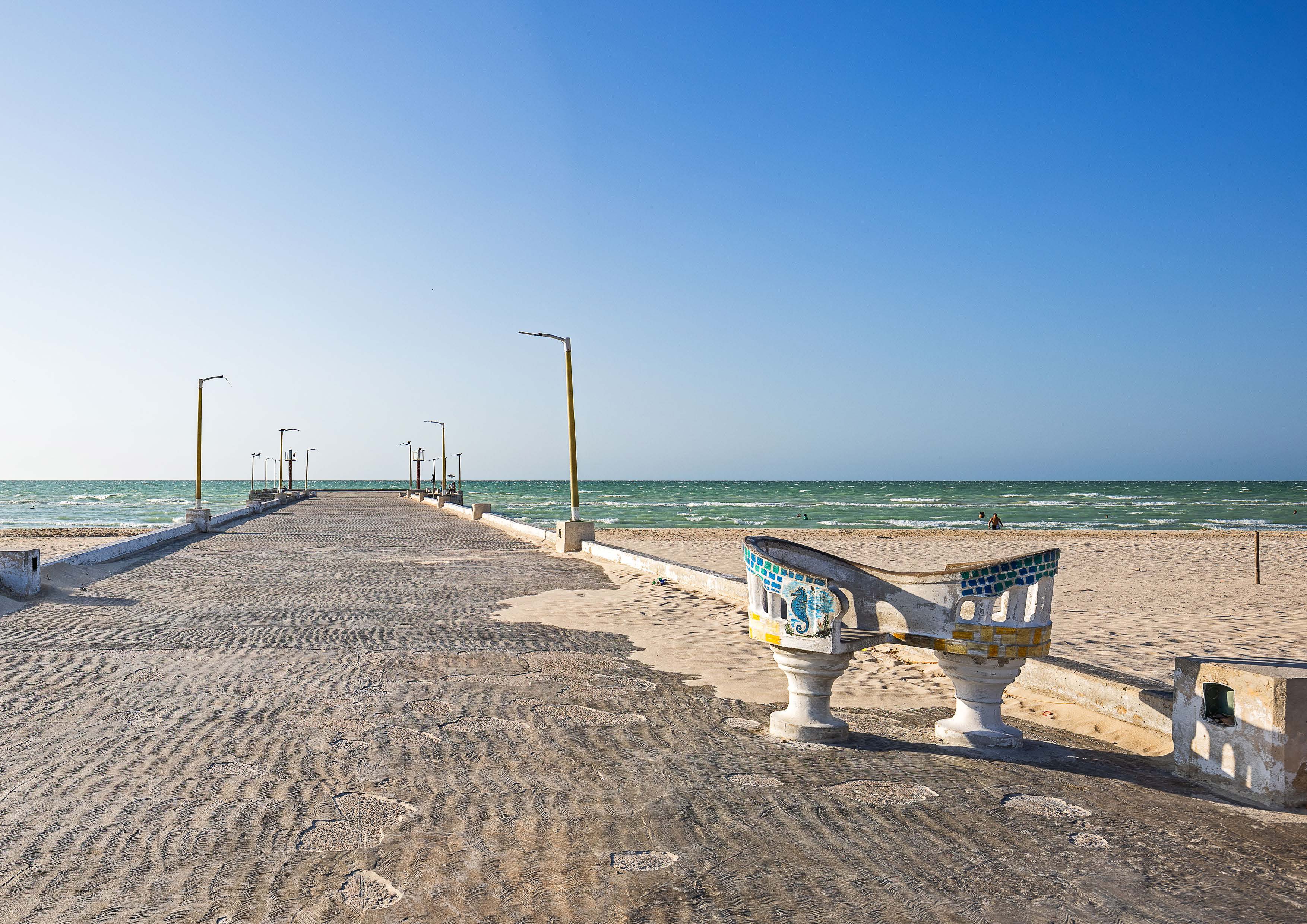 A bench facing the sea at the end of a stone pier under a clear blue sky.