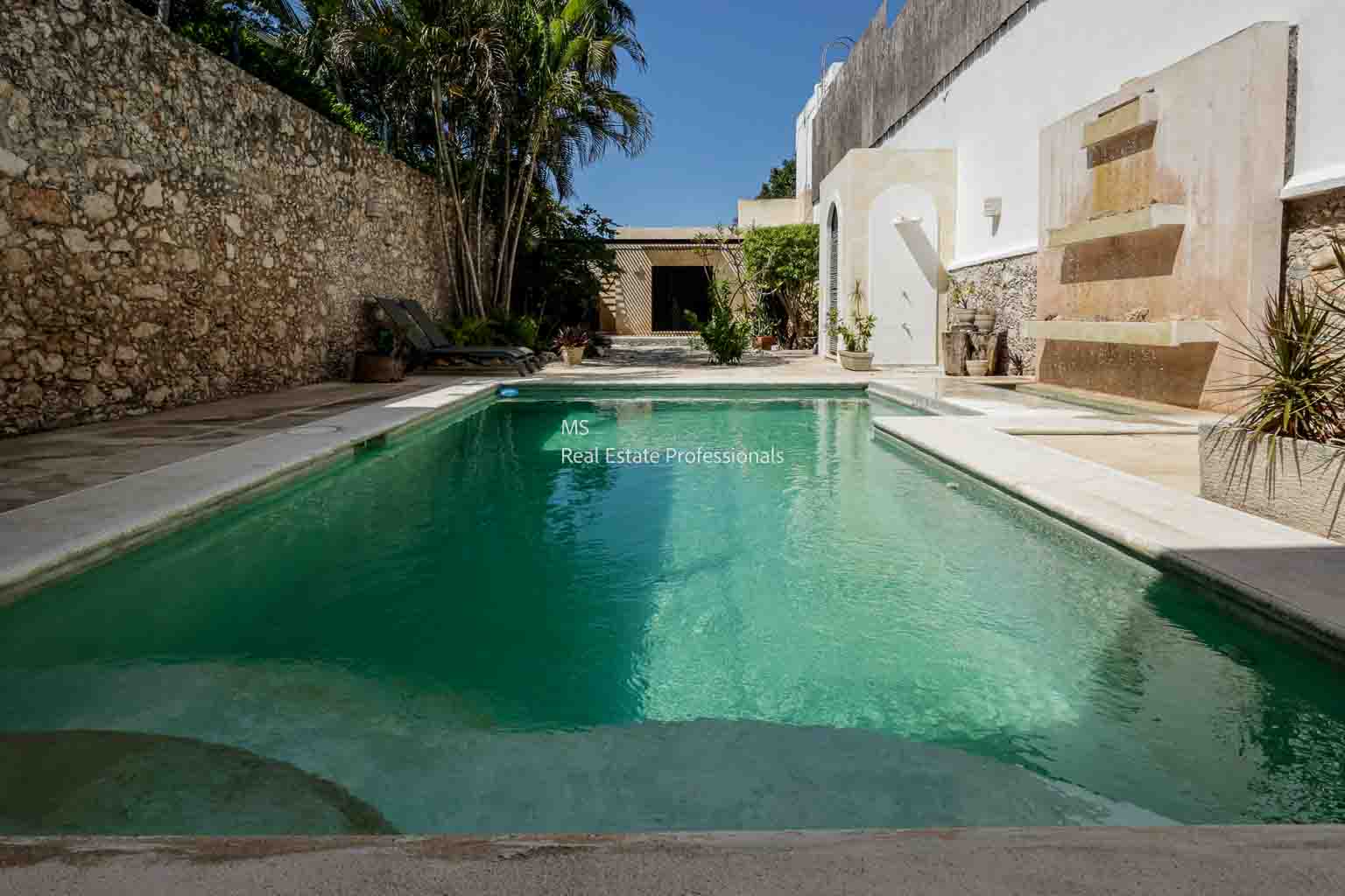 A serene backyard with a clear swimming pool surrounded by stone walls and greenery.