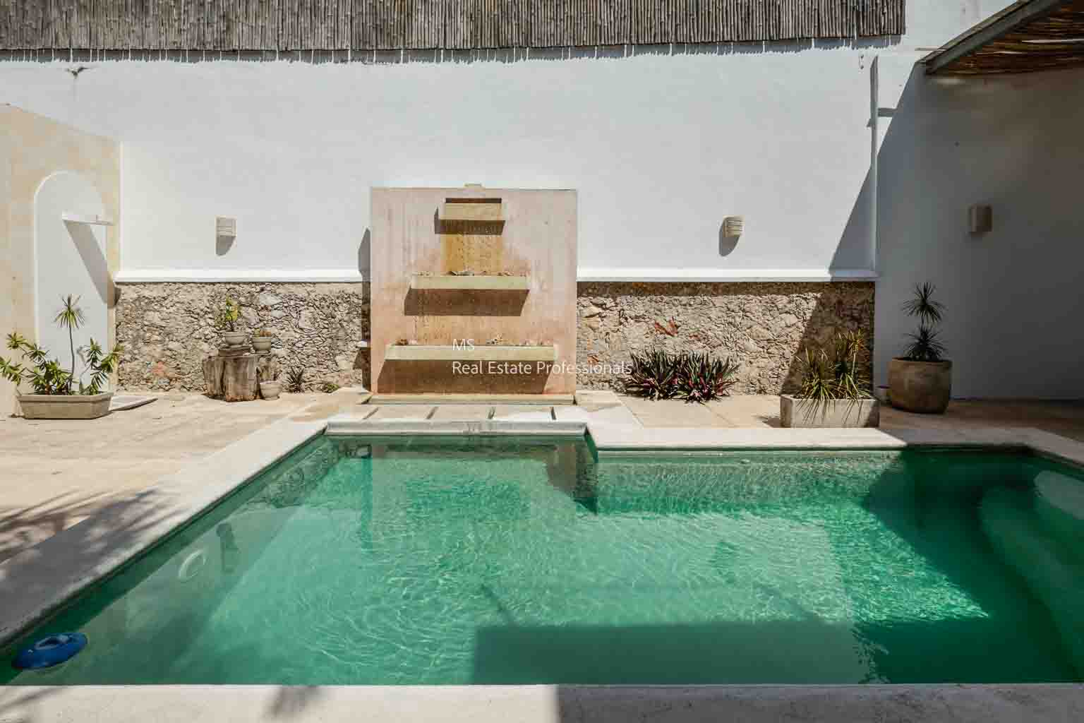 A serene outdoor pool with a stone fountain against a white wall.
