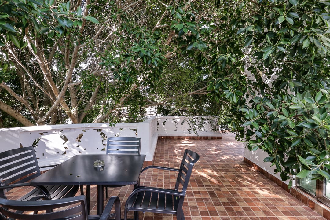 Outdoor patio with a piano and black chairs under leafy trees.