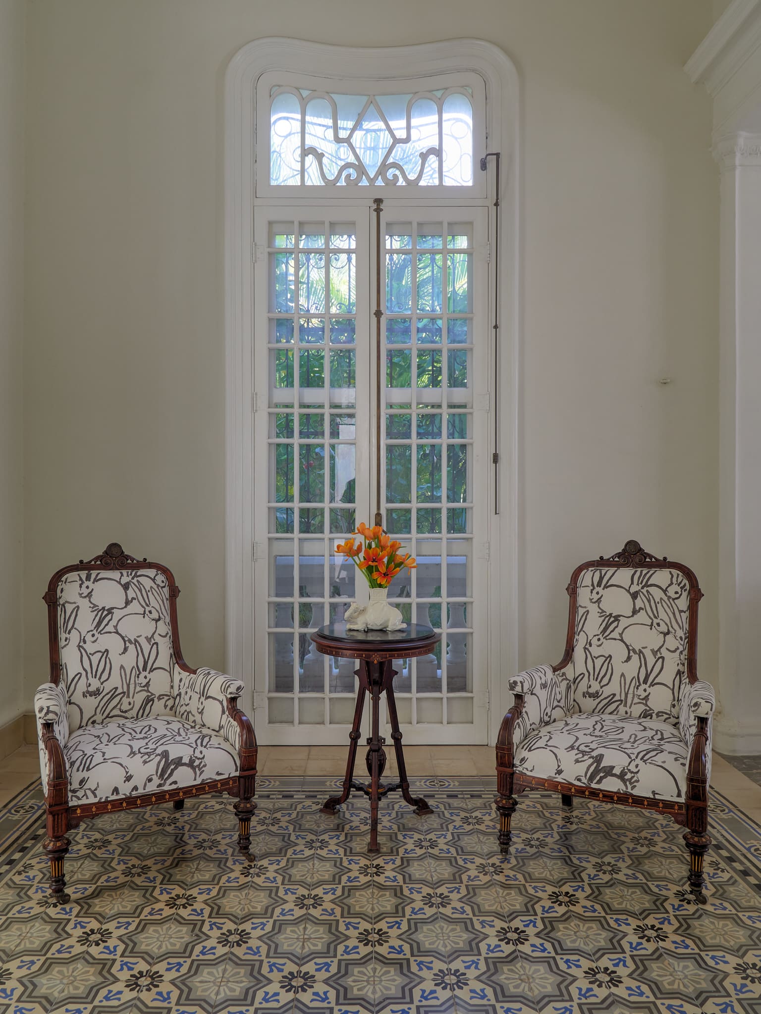 Elegant vintage armchairs flanking a small table with flowers by a large window.