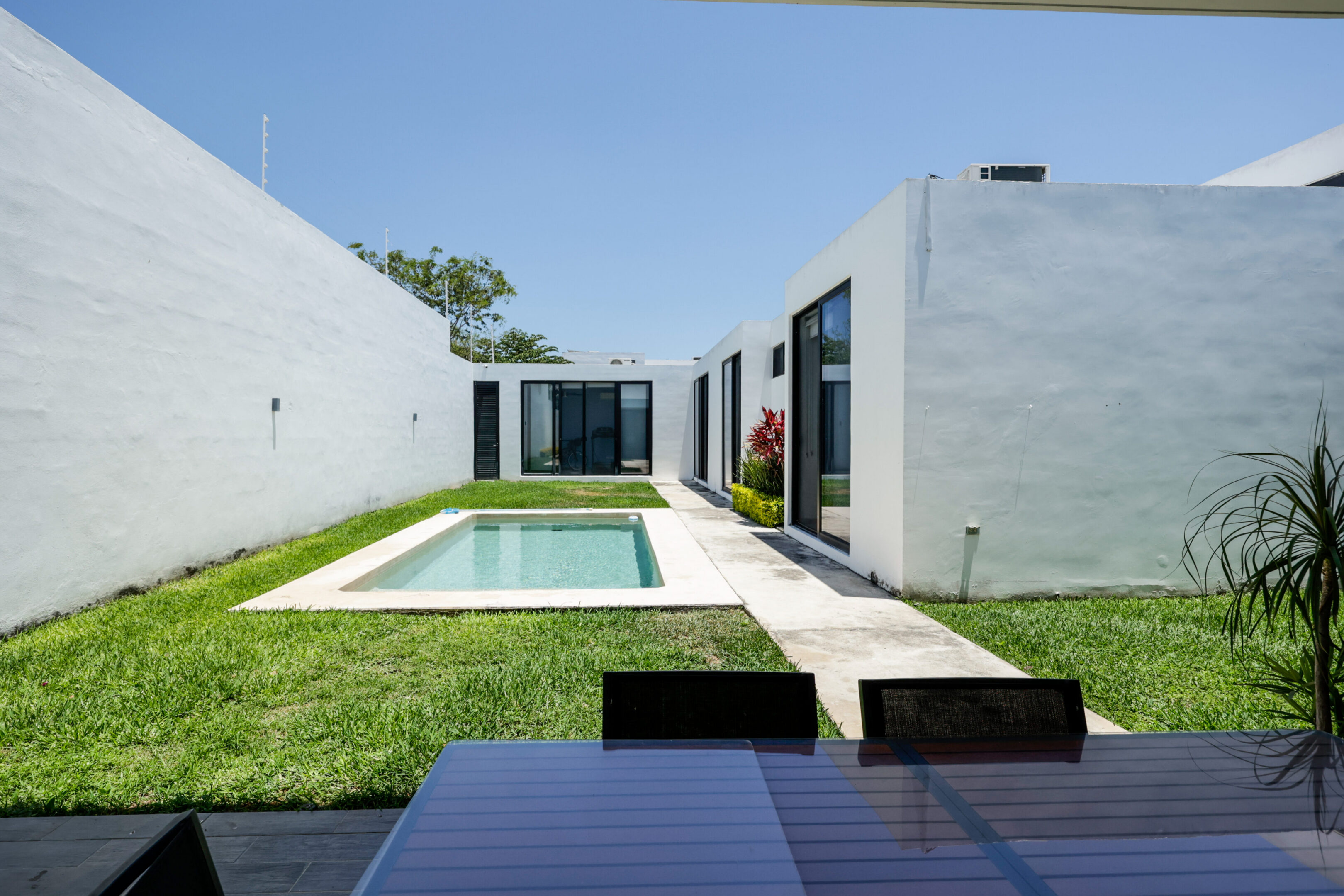 Modern white house with a rectangular pool and glass doors under a clear blue sky.