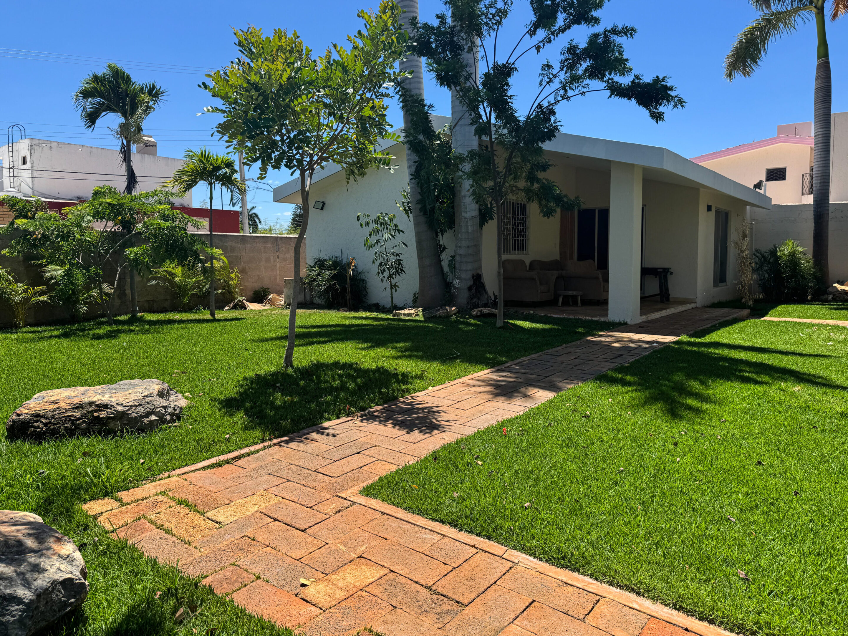 Sunlit backyard with green lawn and brick pathway beside a house.